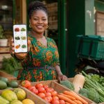 Entrepreneure camerounaise derrière des cagettes de fruits et légumes frais, montrant sa boutique en ligne sur son smartphone